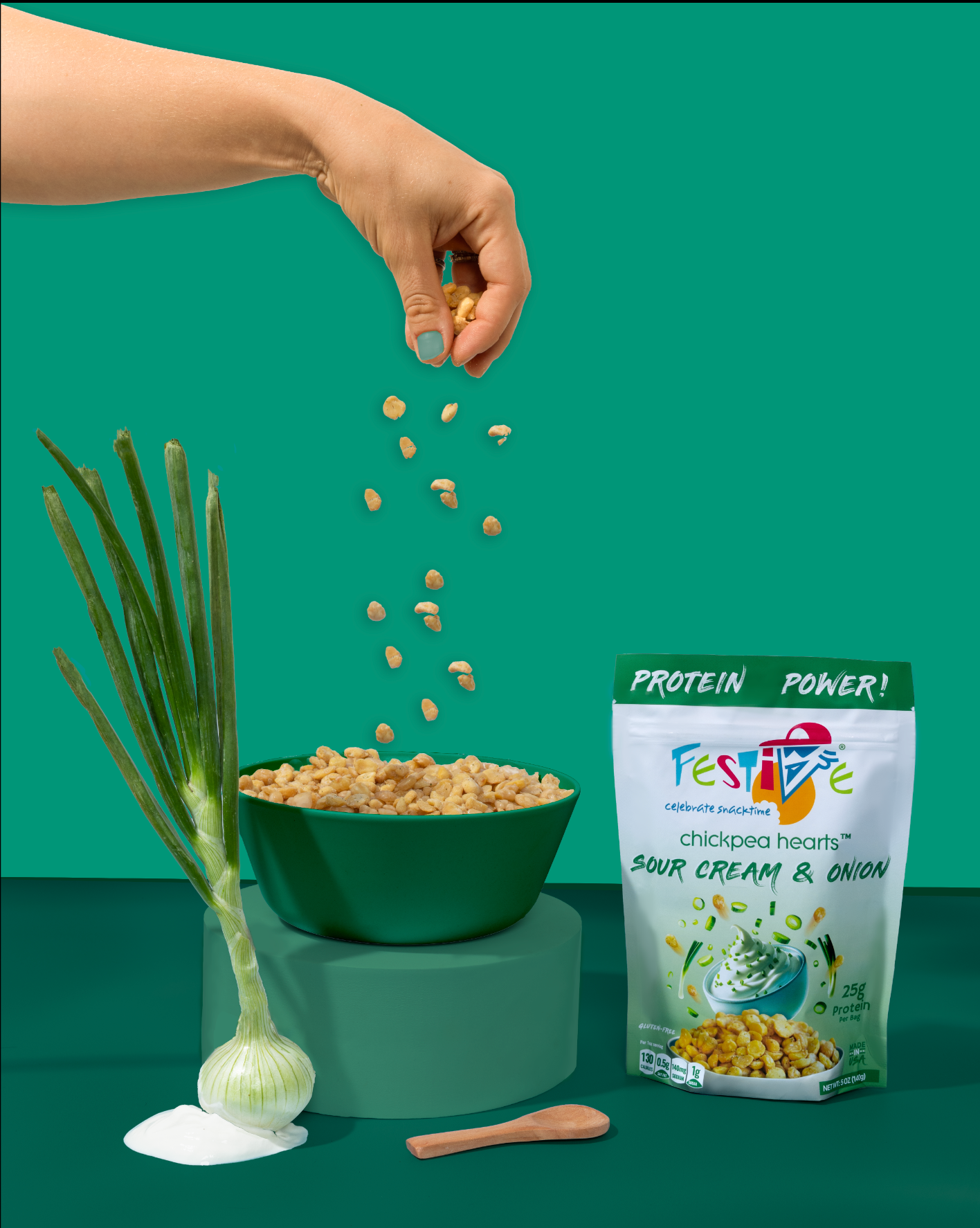 Hand pouring chickpeas into a bowl with a green background, featuring a package of Festive Chickpea Hearts Sour Cream & Onion flavor.
