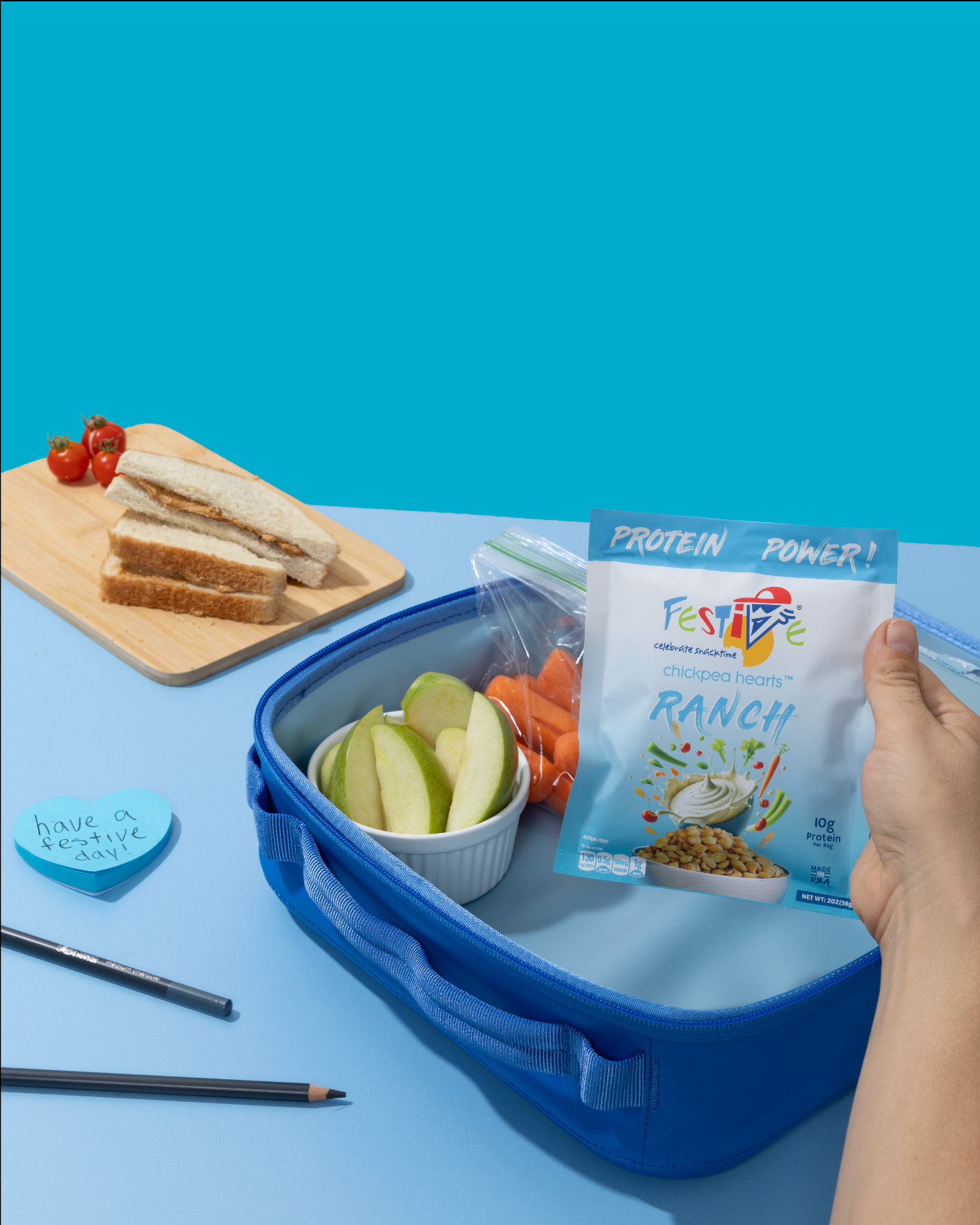 Blue lunch box with snacks and a hand holding a packet of 'Festive' ranch chickpeas on a blue background.