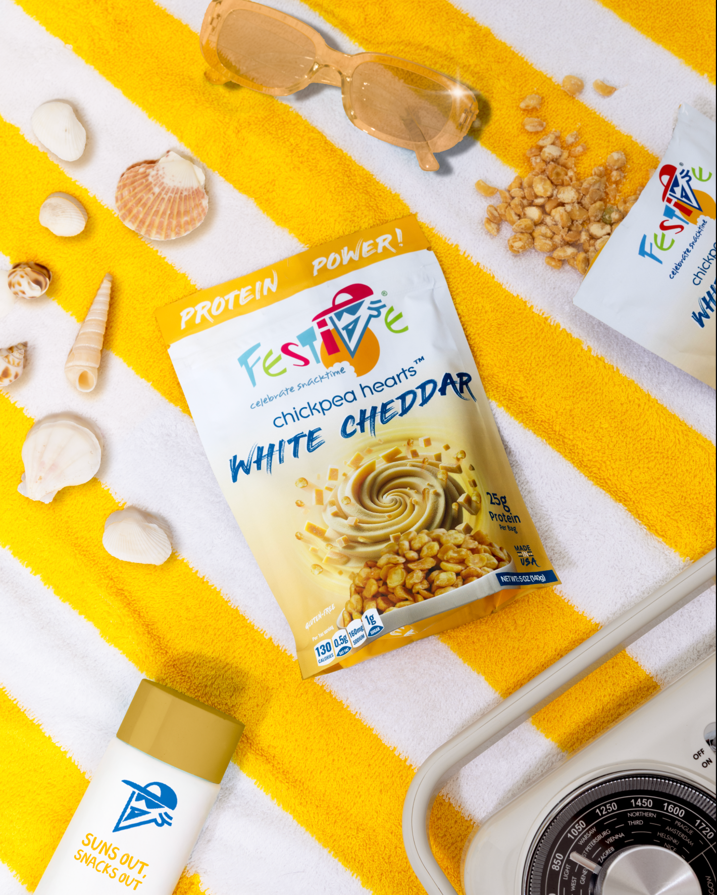Festive chickpea hearts white cheddar package on a yellow and white striped towel with sunglasses and snacks.